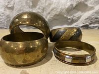 Set of four brass bangles arranged on a surface with a white textured wall in the background, showing variations in design and finish.