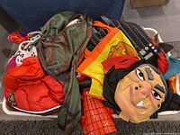 Large plastic bin filled with various children's Halloween costumes and accessories including witch mask, camouflage, red padded costume, spider-man sleeve, yellow pumpkin cape.