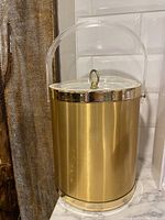 Front angled view of gold-tone metal ice bucket with clear plastic handle and lid with golden knob.