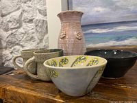 Five pottery pieces including pink vase, two beige mugs, decorated bowl, and dark blue bowl on wooden surface