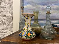 Four vases and glass decanter arranged on a wooden surface with a painting in background. Includes a colorful hand-painted ceramic vase, two greenish glass vases, and a clear glass decanter.