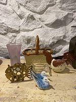 Photo showing small basket with wicker handle, ceramic rooster creamer, fish-shaped trivet, blue and white ceramic birds, and pink glass vase on a shelf against a stone wall.