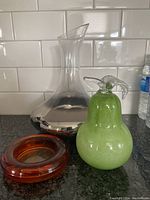 Lot of three vintage glass items: clear glass decanter with silver base, green glass pear figurine, and orange glass dish all displayed on kitchen counter with white tile background.
