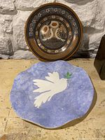 Two pottery serving trays shown together, one round with bird and floral patterns, one irregular shaped with white dove on blue background
