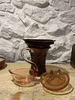 Photo of signed pottery vase with a mottled brown and tan glaze, a peach-colored glass swan figurine, and a matching glass ashtray on a shelf with stone wall background.