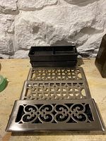 Three metal vent registers laid out flat next to three black plastic vent frames on a wooden surface.