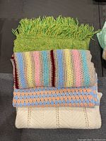 Four stacked vintage blankets showing various crochet and knit patterns including fringed green, multi-color stripes, geometric peach and blue pattern, and cream knit texture.