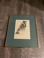 Framed charcoal drawing featuring a male torso holding a rod, signed at bottom right, shown on wooden floor