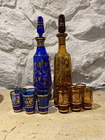 Both blue and amber vintage glassware sets, including decanters with stoppers and matching shot glasses with detailed gold decoration.