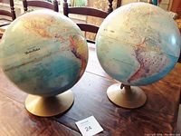 Photo shows two vintage globes side by side on a wooden table with chairs in background.