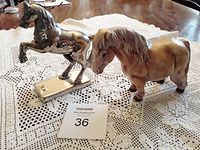 Photo showing the chrome rearing horse bank standing on its rectangular base and the Shetland pony bank side by side on a crochet lace tablecloth.