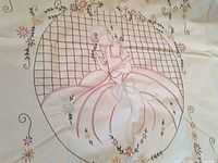 Close-up photo of hand embroidery showing a seated lady in a hoop skirt dress with puffed sleeves and bonnet surrounded by a lattice circle and florals.