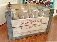 Front angled view of the wooden dairy crate showing stencil 'SILVERWOOD'S 10 LONDON 54' with 12 empty clear glass milk bottles inside