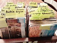 Photo showing multiple stacks of vintage postcards with country name labels including Austria, Belgium, Germany, Holland, India, Israel, Japan, Morocco, Spain, Turkey, U.S.A., and others.