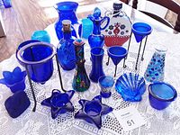 Wide view of entire lot on lace tablecloth showing cobalt blue vintage glass items including decanters, vases, candle holders, and decorative pieces.