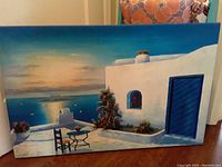 Frontal view of the oil painting showing white buildings, blue door, terrace with table and chairs, pot plants, and sea with sunset sky.