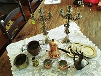 Overview of entire lot showing brass and copper items on lace tablecloth including candelabras, coffee pot with creamer and sugar bowl, brass plates, planters, and additional containers.