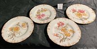 Four antique luncheon plates arranged side by side on dark fabric background showing floral patterns and gold-tone decorated borders.