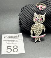 Photo shows the owl brooch and ring resting on a black ridged fabric background alongside a lot number card for auction.