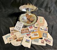 Photo showing silver plated clam-shaped trinket box open with assorted coins inside and postage stamps laid out below on a dark surface.