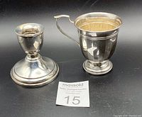 A front view photo of a sterling silver weighted candlestick and a lidded sterling silver cup side by side on black background with auction lot number visible.