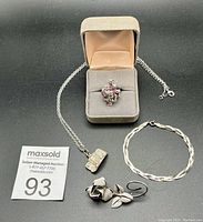 Photo showing all four items together: pinky ring with garnets in a jewelry box, rectangular pendant on chain, tri-coloured bracelet, and rose-shaped brooch.