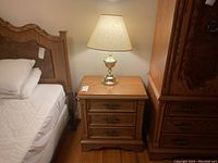 Oak nightstand with brass lamp on top next to bed, showing full piece and context with mattress and pillows.