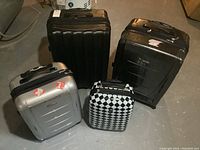 Four suitcases arranged on floor. Large black London suitcase, medium black Samsonite, small silver Samsonite, and black/white diamond Heys suitcase visible.
