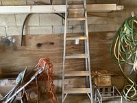 Aluminum step ladder standing in a garage against a cinder block wall, next to coiled garden hoses and other garage tools.
