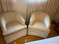 Pair of beige velvet boudoir chairs side by side on hardwood floor near window with curtains