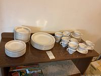 Photo showing stacked white dinner plates, side plates, cups and saucers on a wooden table.