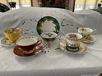 Front view of five cups and saucers showing different color schemes and intricate patterns, displayed on white embroidered fabric.