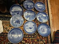 Overview photo showing 10 of the blue and white collector plates laid out on a carpet, depicting various Christmas and winter scenes including cottages, birds, carolers, and harbor imagery.