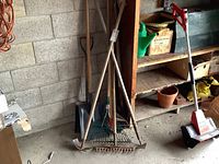 Photo showing assortment of garden tools standing upright including rakes, shovels, and hoes arranged against a wall.