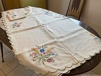 Full view of cream linen tablecloth spread on rectangular table with 4 embroidered floral corner designs including wheat sheaf and blue cornflowers. 10 matching napkins folded on table.