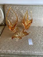 Three amber-colored MCM art glass sculptures displayed on a fabric-covered couch, showing the set and relative sizes.