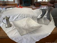 Wide view of large square aluminum bowl, two matching vases, and pewter stein displayed on a white embroidered cloth on a wooden table.
