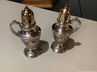 Close-up photo of pair of silver salt and pepper shakers with gold wash lids showing intricate detailing and ornate handles.