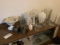 Photo showing a collection of assorted glass and ceramic vases and decorative items on a wooden surface.