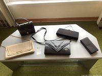 Four vintage bags displayed on a white table including black structured handbag with top handle, black leather shoulder bag, beige cloth clutch, and black wallet