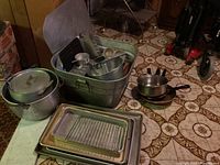 Full view of metal wash tub containing pots and kitchen tools, surrounded by aluminum pots and stacked baking trays on floor.