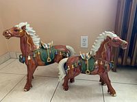 Two ceramic carnival horse figurines showing side view and decorative saddles with tassels, set on tile floor near wall outlet.