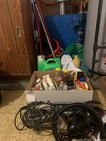 Photo showing cardboard box with assorted cleaning products including liquids and sprays, placed on floor in basement near blue furnace.