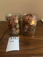 Two glass jars filled with mixed Canadian and American pennies, displayed on a wooden surface with a lot tag.