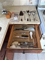 View of kitchen counter with knife block holding steak knives, large serving utensils laid out on counter, and open drawer organizer containing various cutlery and utensils.