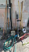 Several long-handled garden tools including rakes, shovels, a green hose coiled on ground, a sledge hammer, garden saw, and hedge trimmer leaning against exterior wall
