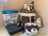Photo showing the raised toilet seat box, white shower chair, handheld massager, blood pressure monitor, and foot bath on tiled floor in corner.