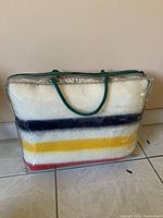 Throw blanket packed in a clear plastic bag with green handles showing the multicolor stripes on cream base.