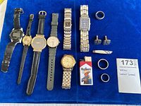 Display of men’s wristwatches with different styles and bands, a few rings, a Marlboro lighter, and a set of cuff links and tie clip on blue velvet surface.