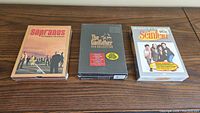 Three unopened DVD sets on a wooden surface: The Sopranos Complete Third Season, The Godfather Trilogy, and Seinfeld Season 8.